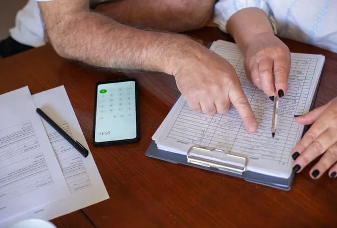 Photograph of two people at a wooden table reviewing a printed grid of plan details on a clipboard, one pointing at a row while the other uses a calculator app on a phone beside unsigned forms.