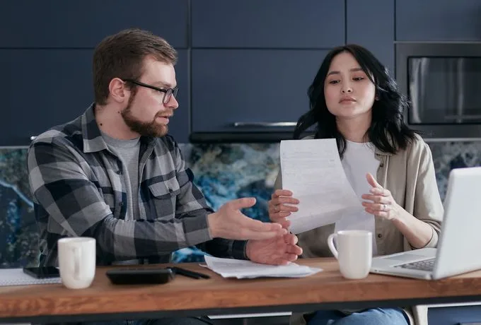 A man and a woman at a kitchen counter talking over a printed letter, with a laptop and two mugs in front of them.