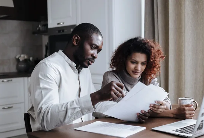 A couple sits at a kitchen table reviewing printed papers together, with an open laptop and a coffee mug in front of them.
