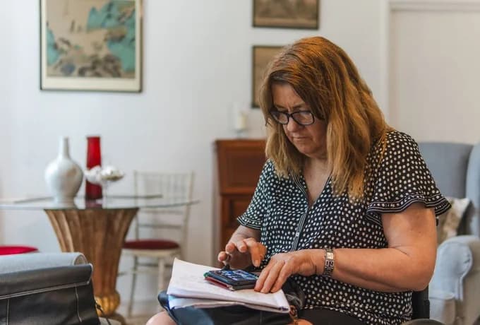 A woman wearing glasses sits in a living room, reviewing papers in her lap while holding a smartphone.