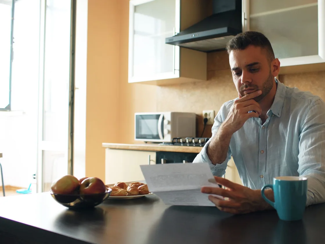 A man reading a letter at a sunlit kitchen table.
