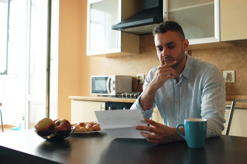 A man reading a letter at a sunlit kitchen table.